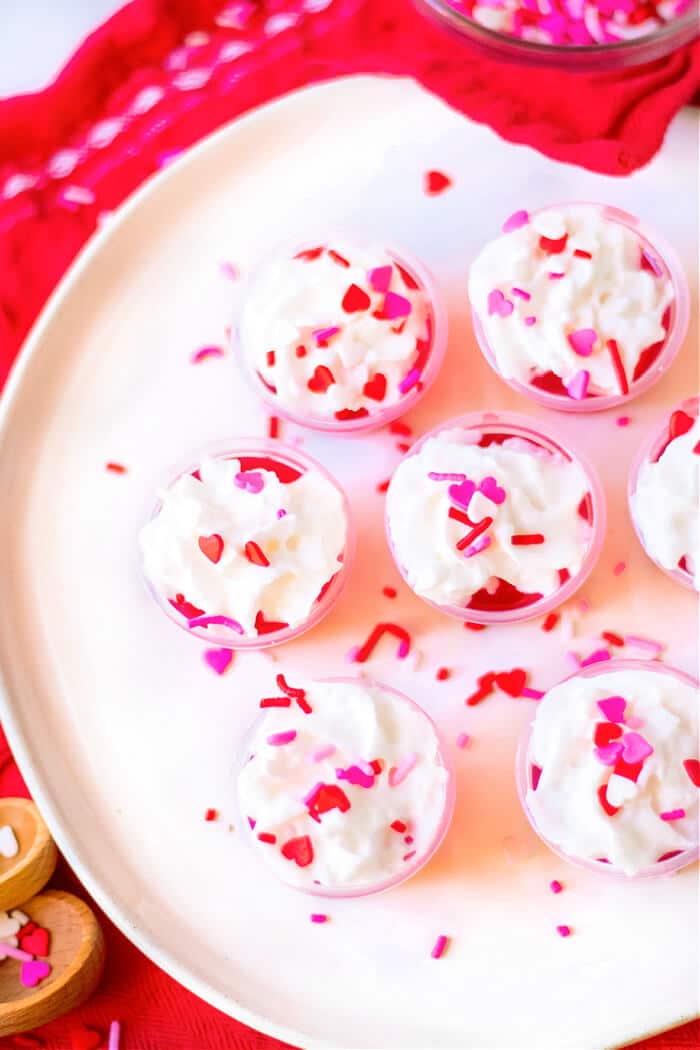 Valentine’s jello shots are a festive layered shot of raspberry and strawberry jello topped with whipped cream & sprinkles! RECIPE FOR VALENTINE'S JELLO SHOTS