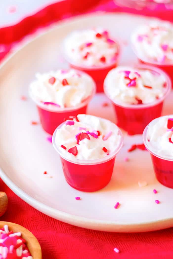 Valentine’s jello shots are a festive layered shot of raspberry and strawberry jello topped with whipped cream & sprinkles! VALENTINE'S JELLO SHOTS RECIPE